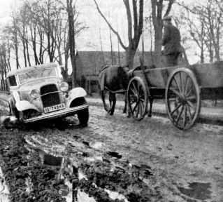 Das schwarzweiß Foto zeigt eine Landstraße in Niedersachsen um 1925, worauf ein im Matsch eingesunkenes Auto und eine Kutsche zu sehen ist.